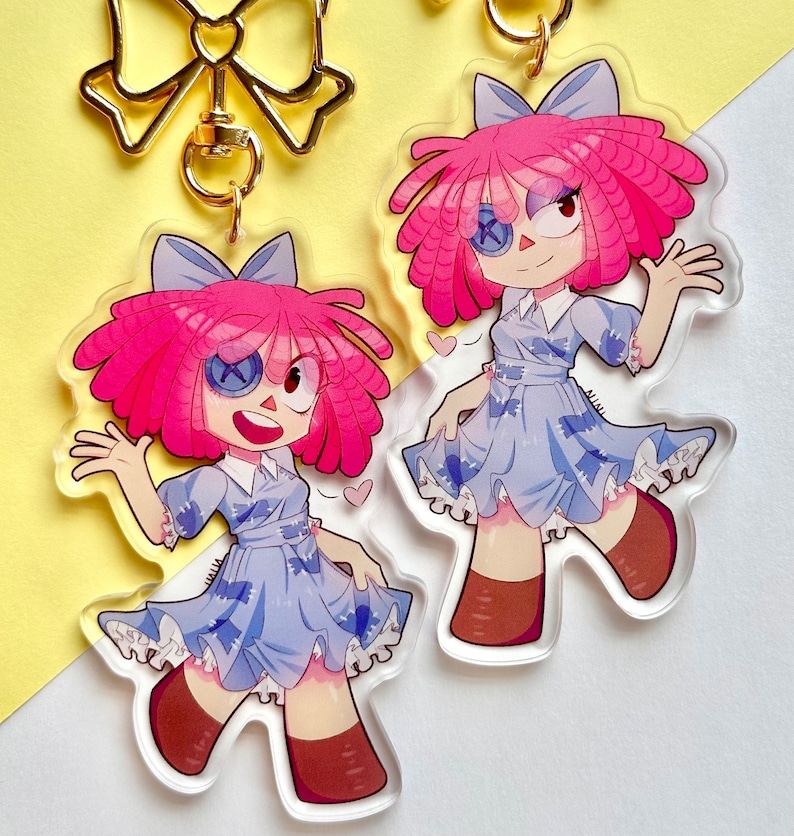 Puede incluir: Dos llaveros de acr&iacute;lico transparente con un dise&ntilde;o de mu&ntilde;eca de dibujos animados. La mu&ntilde;eca tiene pelo rosa, un vestido azul y botas marrones. Los llaveros est&aacute;n sujetos a cierres dorados en forma de lazo. El fondo es una mezcla de amarillo y blanco.