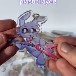 Puede incluir: Un dije acr&iacute;lico transparente con un conejo de dibujos animados tocando una guitarra rosa. El conejo es morado con una pajarita roja. El dije tiene una capa de pl&aacute;stico removible. El texto en la imagen dice "Has a removable plastic layer!" y "May seemed scratched but once removed it will be smooth!"
