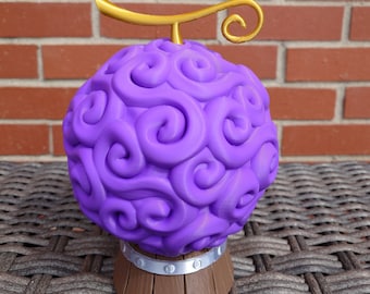 May include: A purple and gold 3D printed figurine of a devil fruit, a spherical object with swirling patterns, on a brown wooden base.