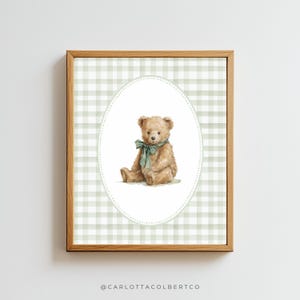 May include: A framed print featuring a watercolor illustration of a teddy bear with a green bow. The bear is centered within an oval frame against a light green and white gingham pattern. The frame is made of wood.