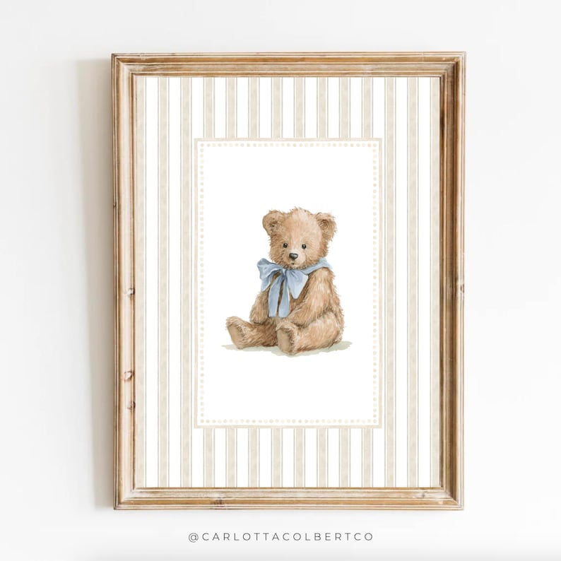 May include: A framed print featuring a watercolor illustration of a teddy bear with a blue ribbon. The bear is light brown and sits against a white background with a decorative border. The frame is a light brown color with a striped background.