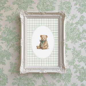 May include: A framed print featuring a watercolor illustration of a teddy bear with a teal bow. The bear is set against a light green and white gingham pattern within an oval frame. The white ornate frame is mounted on a floral wallpaper.