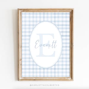 May include: A framed print with a light blue and white gingham pattern. The central oval displays the name "Everett" in a script font, with a large letter "E" behind it. The frame is a light brown colour.