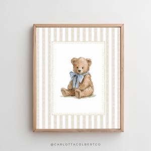 May include: A framed print featuring a watercolor illustration of a teddy bear with a blue bow. The bear is set against a striped background in beige and white, within a decorative border. The frame is made of light-colored wood.