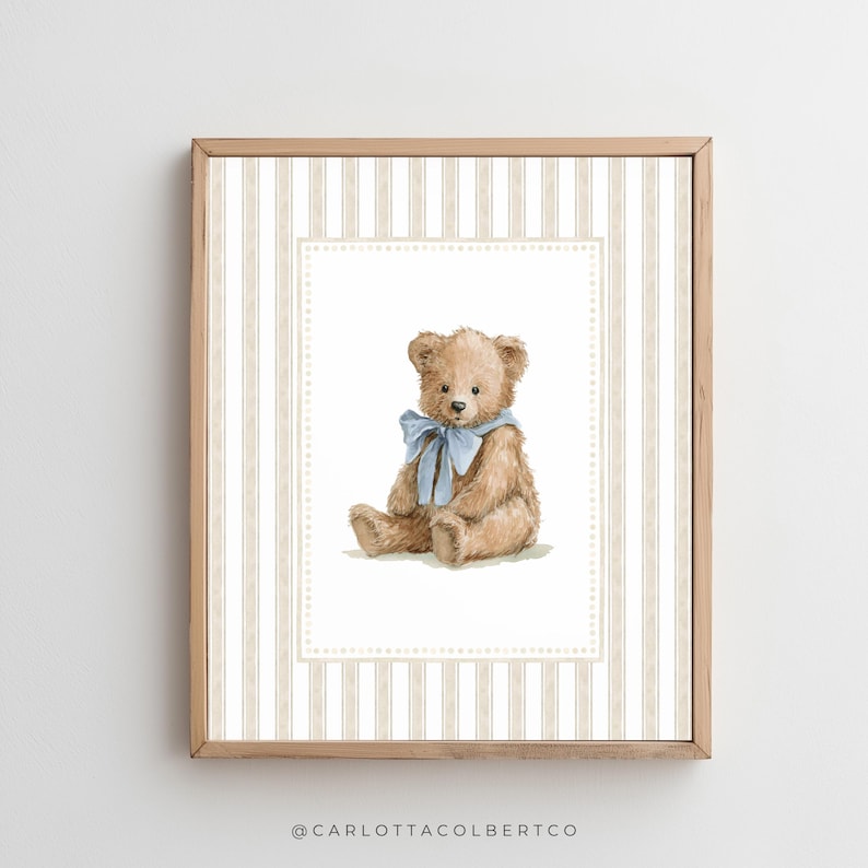 May include: A framed print featuring a watercolor illustration of a teddy bear with a blue bow. The bear is set against a striped background in beige and white, within a decorative border. The frame is made of light-colored wood.