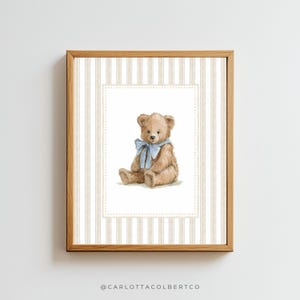 May include: A framed print featuring a watercolor illustration of a teddy bear. The bear is light brown with a blue ribbon around its neck. The background has a striped pattern in beige and white.