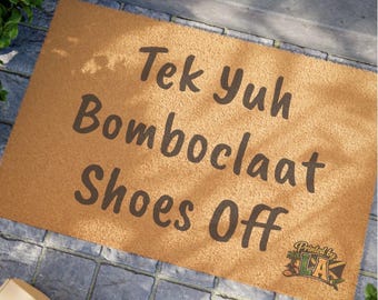 Pode incluir: Um capacho retangular com o texto "Tek Yuh Bomboclaat Shoes Off" em letras marrons escuras. O tapete é de cor marrom claro e tem uma superfície texturizada. As palavras "Printed by LA" estão no canto inferior direito.