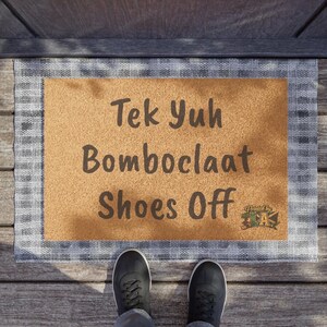Pode incluir: Um tapete retangular com o texto "Tek Yuh Bomboclaat Shoes Off" em castanho escuro. O tapete tem um centro castanho claro e uma borda xadrez cinza e branca. Sapatos pretos s&atilde;o vis&iacute;veis na parte inferior.