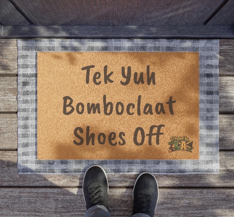 Pode incluir: Um tapete retangular com o texto "Tek Yuh Bomboclaat Shoes Off" em castanho escuro. O tapete tem um centro castanho claro e uma borda xadrez cinza e branca. Sapatos pretos s&atilde;o vis&iacute;veis na parte inferior.