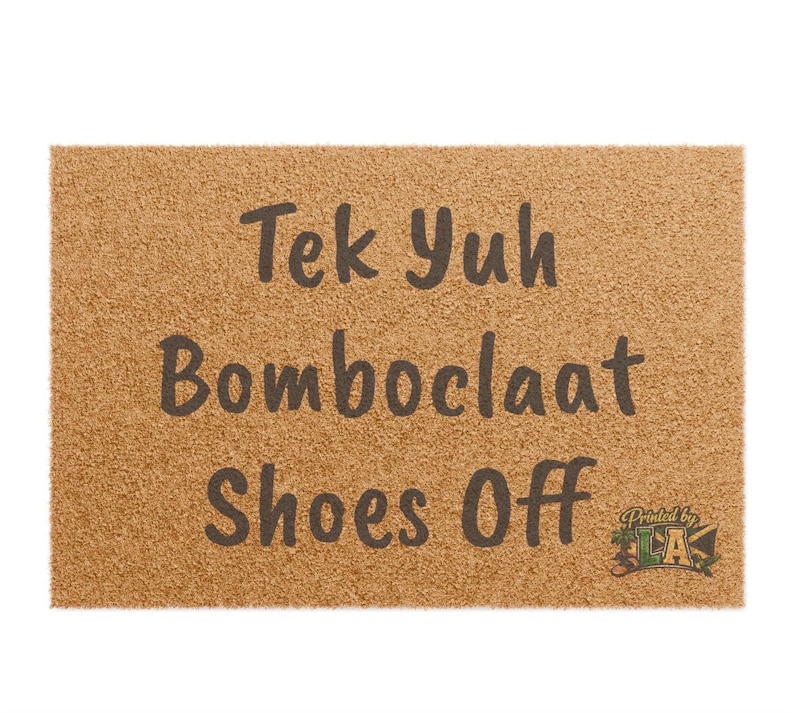 Pode incluir: Um capacho retangular com uma superf&iacute;cie texturizada castanho-clara. O tapete apresenta o texto "Tek Yuh Bomboclaat Shoes Off" em letras castanho-escuras. As palavras s&atilde;o empilhadas verticalmente. Um logotipo no canto inferior direito.