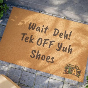Pode incluir: Um capacho retangular castanho claro com o texto "Wait Deh! Tek OFF Yuh Shoes" em castanho escuro. O capacho tem um logotipo no canto inferior direito que diz "Printed by LA".