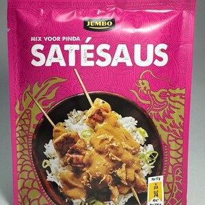 Pode incluir: Um pacote rosa e dourado de mistura para molho satay. O pacote apresenta uma foto de um espeto de frango satay com arroz e um design de dragão. O texto no pacote diz "JUMBO MIX VOOR PINDA SATÉSAUS".