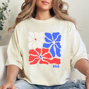 May include: A cream-colored t-shirt with a graphic of three stylized flowers in red, white, and blue. The flowers are arranged in a square shape and the word "USA" is printed below them.