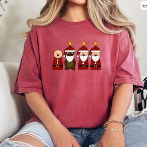 May include: A crimson red t-shirt featuring a graphic of five cartoon figures. The figures are wearing Santa hats and have flames on top of their heads. The figure in the centre is brown and the other four are red.