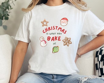 Könnte beinhalten: Weißes T-Shirt mit dem Text "Christmas is what you bake it!" und Illustrationen von Lebkuchen, darunter ein Schneemann, ein Stern, eine Schneeflocke, der Weihnachtsmann und ein grüner Christbaumschmuck.