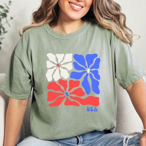 May include: A green t-shirt with a graphic of three stylized flowers in red, white, and blue. The flowers are arranged in a square shape and the word "USA" is printed below them.