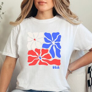 May include: White t-shirt with a graphic of three stylized flowers in red, white, and blue. The flowers are arranged in a square shape. The text "USA" is printed below the flowers.
