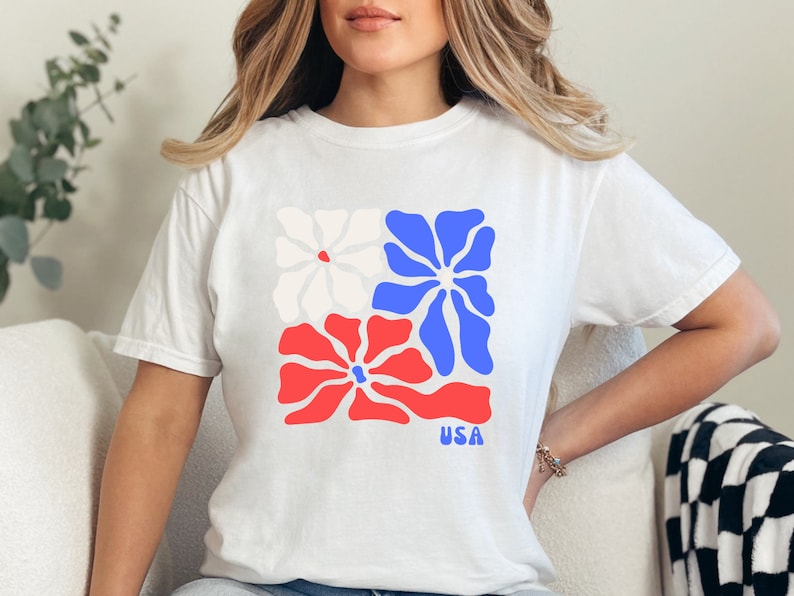 May include: White t-shirt with a graphic of three stylized flowers in red, white, and blue. The flowers are arranged in a square shape. The text "USA" is printed below the flowers.