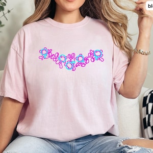 Puede incluir: Una camiseta rosa con un dise&ntilde;o gr&aacute;fico de flores azules y rosas. Las flores est&aacute;n dispuestas en una l&iacute;nea a trav&eacute;s del pecho de la camiseta.