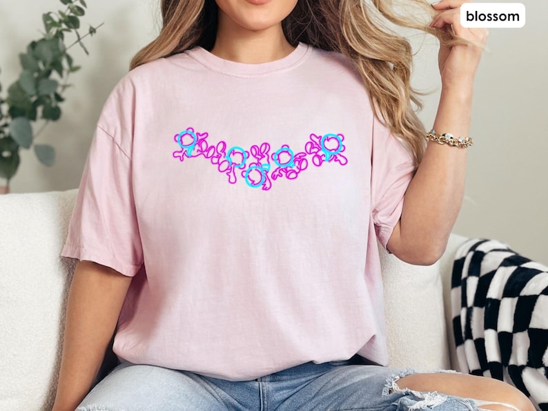 Puede incluir: Una camiseta rosa con un dise&ntilde;o gr&aacute;fico de flores azules y rosas. Las flores est&aacute;n dispuestas en una l&iacute;nea a trav&eacute;s del pecho de la camiseta.