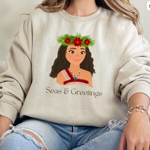 May include: A beige sweatshirt featuring a cartoon illustration of a woman wearing a flower crown and the text "Seas & Greetings".