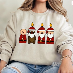 May include: A beige sweatshirt with a Christmas-themed design featuring four cartoon figures. The figures are a snowman, a reindeer, and two Santas with candles on their heads. The design is in red, brown, and yellow.