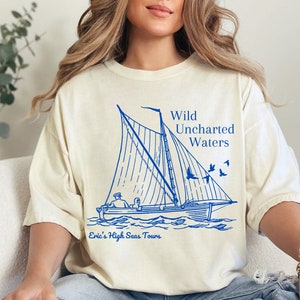 Puede incluir: Una camiseta blanca con una ilustración azul de un velero con el texto "Wild Uncharted Waters" y "Eric's High Seas Tours" impreso en ella.