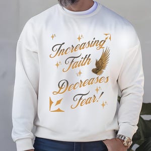 May include: Cream-colored sweatshirt with the text "Increasing Faith Decreases Fear!" in a stylish font. The design includes a gold-colored wing and star accents. The sweatshirt is a comfortable, casual style.