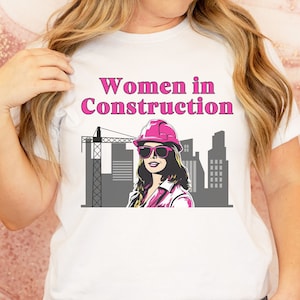 May include: White t-shirt with a pink graphic of a woman wearing a pink hard hat and sunglasses. The graphic also includes a gray construction crane and buildings. The text "Women in Construction" is printed in pink on the shirt.