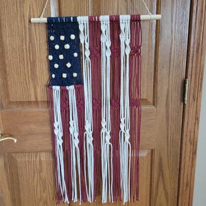 May include: A macrame wall hanging in the shape of the American flag. The flag is made with red, white, and blue yarn and has wooden beads for the stars.