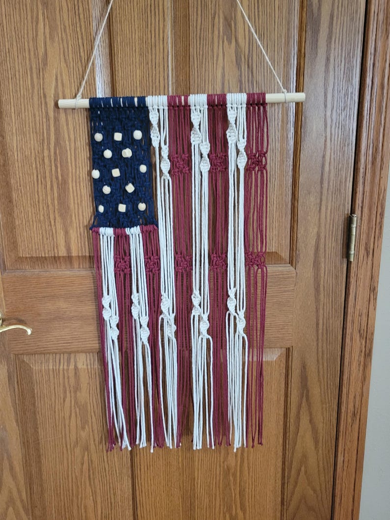 May include: A macrame wall hanging in the shape of the American flag. The flag is made with red, white, and blue yarn and has wooden beads for the stars.
