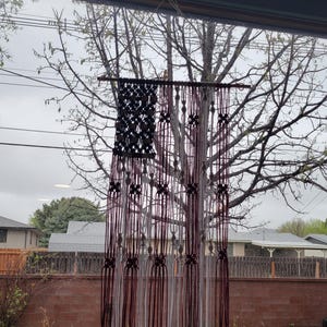 May include: A red, white and blue macrame wall hanging in the shape of an American flag. The hanging is made with white and burgundy yarn and is hanging in front of a window.