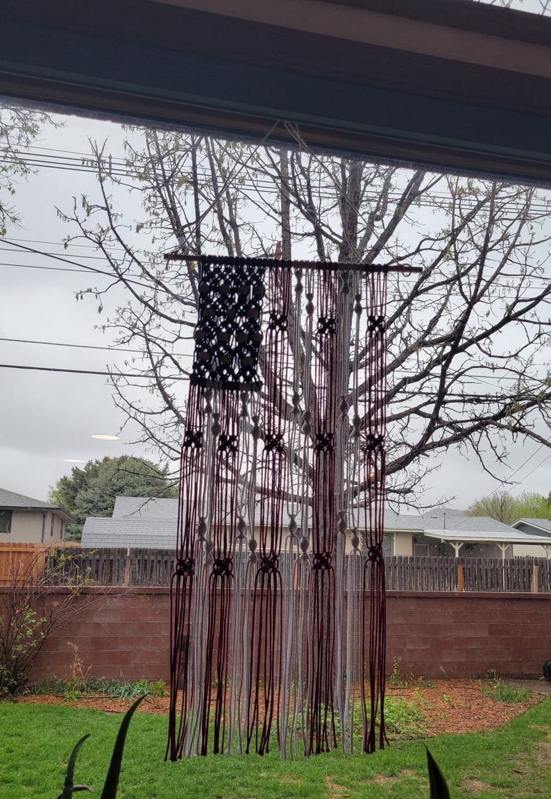 May include: A red, white and blue macrame wall hanging in the shape of an American flag. The hanging is made with white and burgundy yarn and is hanging in front of a window.