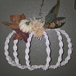 May include: A wall hanging shaped like a pumpkin, constructed from white macrame cord in a twisted pattern. The top is decorated with a small white pumpkin, beige silk flowers, brown autumn leaves, green foliage, and small dried berry clusters.