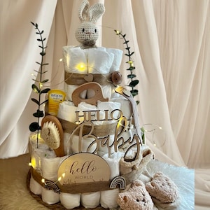 Könnte beinhalten: Eine dreistöckige Windeltorte, verziert mit einem gehäkelten Hasen, Holzspielzeug und einem Schild mit der Aufschrift "Hello Baby". Die Torte ist mit einem Jutestoffband umwickelt und mit Lichterketten geschmückt. Die Torte steht auf einem geflochtenen Untergrund und ist von einer weißen, flauschigen Oberfläche umgeben.