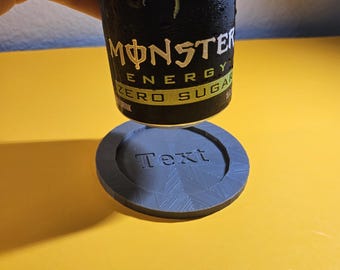Puede incluir: Una lata de bebida energética Monster negra y verde con condensación, sobre un posavasos gris. El posavasos tiene la palabra "Text" en relieve. El fondo es amarillo.