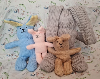 May include: A selection of hand-knitted bunny toys in different colours. There is a large grey bunny, a light blue bunny with yellow ears, a pink bunny, and a tan bunny. The toys are displayed on white fabric with a floral pattern.