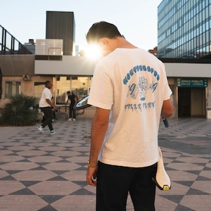 May include: A person wearing a white t-shirt with blue text and a graphic of a cartoon character. The text on the shirt reads "MONSTER LIFE" and "1994" with a blue flower graphic. The person is wearing black jeans and is standing on a checkered floor.