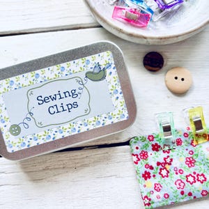May include: A metal tin with the text "Sewing Clips" and a floral design, with sewing clips, buttons, and a fabric swatch. The fabric has a red and white floral pattern. The sewing clips are in various colours.