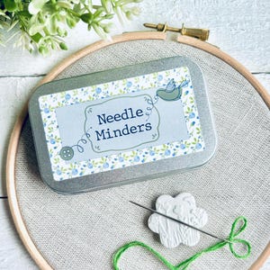 May include: A small tin with a floral design and the text "Needle Minders" on the front. The tin is sitting on a light brown fabric surface. A white ceramic needle minder with a floral design is also visible.