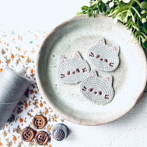 May include: Three grey cat-shaped embroidered patches with brown whiskers and pink ears sit on a speckled ceramic plate. A spool of grey thread, wooden buttons, and a thimble are nearby on floral fabric.