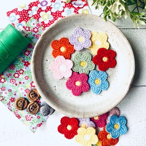 May include: A collection of crocheted flower embellishments in various colours, including pink, blue, green, yellow, and red. The flowers are arranged on a white ceramic plate with a speckled finish.