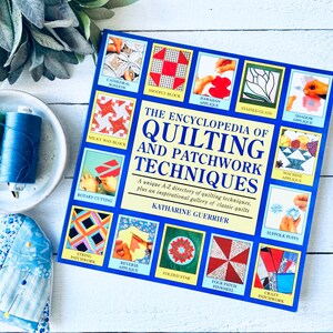 May include: A blue book titled "The Encyclopedia of Quilting and Patchwork Techniques" by Katharine Guerrier. The cover features illustrations of different quilting techniques, including Cathedral Window, Shoo Fly Block, Hawaiian Applique, and String Patchwork.