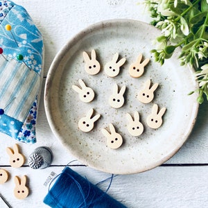 May include: Wooden bunny-shaped buttons with black dot eyes, scattered on a ceramic plate and a white surface. A blue pin cushion, thimble, and blue thread are also visible. The image suggests a sewing or crafting theme, with the buttons as decorative elements.