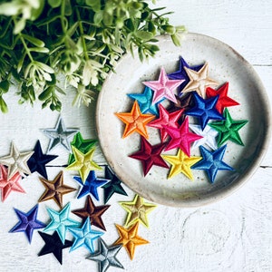 May include: A collection of colourful felt star-shaped patches in various sizes and colours. The stars are arranged in a circle on a white surface.