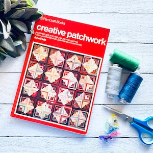 May include: A red book titled "creative patchwork" featuring a colourful quilt pattern on the cover. The book is surrounded by sewing supplies, including spools of thread, scissors, and clips.