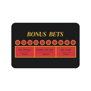 May include: Black rectangular casino-themed mat with the text "BONUS BETS" in gold. It features red boxes with betting options and odds, along with numbered circles. The mat is designed for casino games.