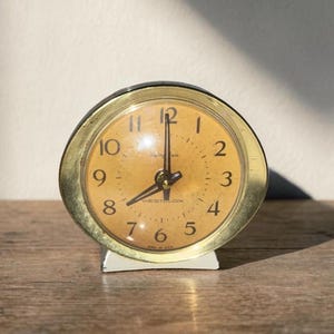 May include: A vintage, round alarm clock with a gold-colored bezel and a cream-colored face. The clock has black numerals and hands, and sits on a white base. The clock is on a wooden surface.