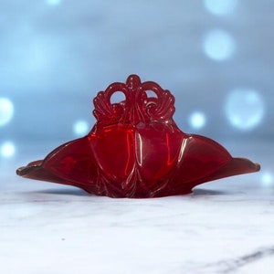 May include: A red glass napkin holder with a decorative design. The holder is shaped like a crown with a pointed top and two curved sides.