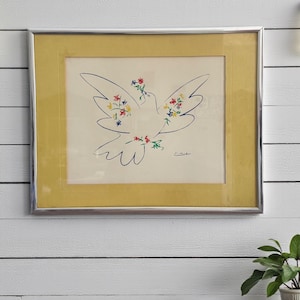 May include: A framed artwork featuring a dove with floral accents. The dove is outlined in blue, with colorful flowers adorning its wings and body. The artwork is set against a yellow background and framed in silver.